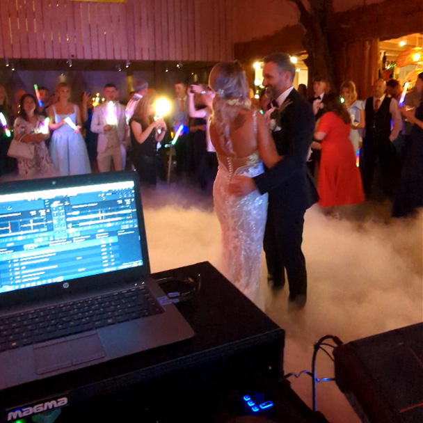 Bride and groom sharing their first dance surrounded by low fog on the dancefloor at a wedding reception while the DJ performs.